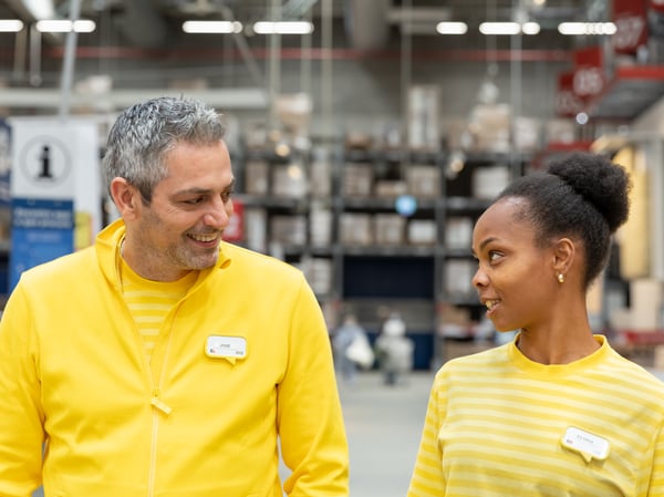 Two IKEA co-workers dressed in yellow work clothes are chatting in the corridor of the self-service warehouse.