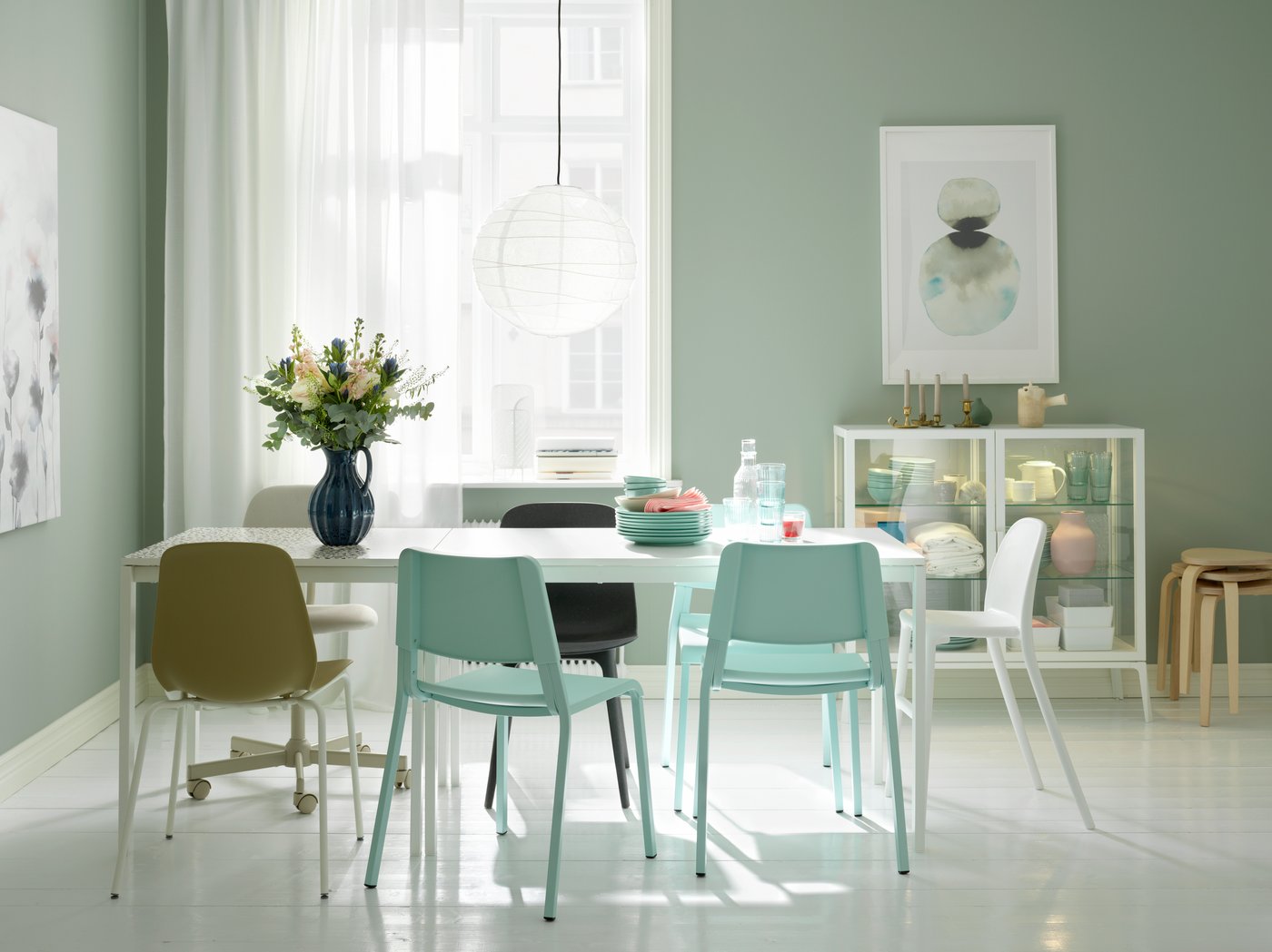 Una zona de comedor formada por dos mesas MELLTORP blancas en línea, una lámpara REGOLIT blanca, sillas variadas y flores en un jarrón.