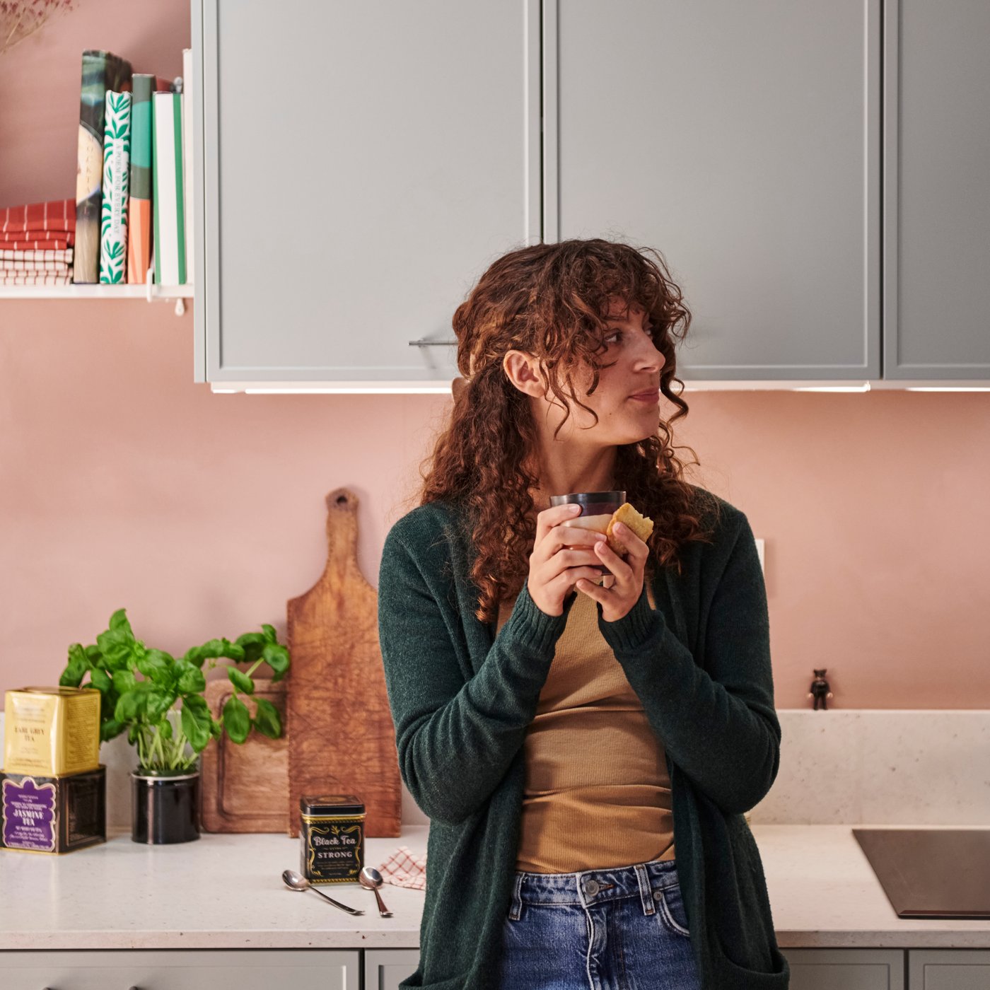 A person holds a tea cup and biscuit in a modern kitchen with ASPUDDEN light grey door fronts and soft pink walls.
