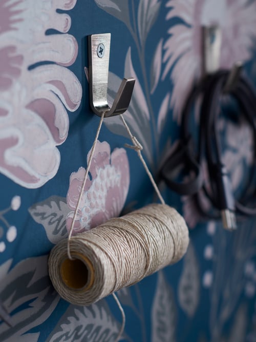 A floral-pattern wall fitted with BLECKA hooks holding a roll of string and connecting cords.