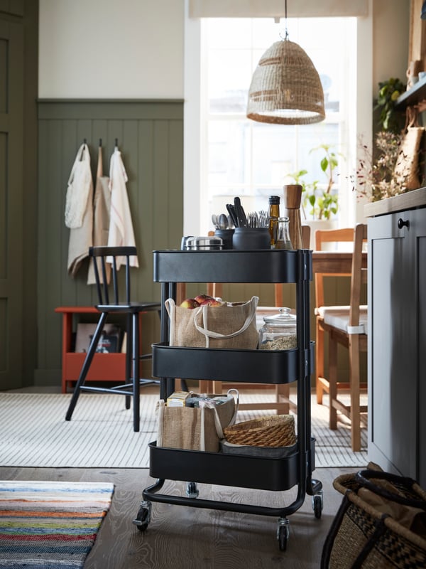 A black RÅSKOG trolley with many things on it, including food and GULLRISMOTT food storage bags, stands near a table.