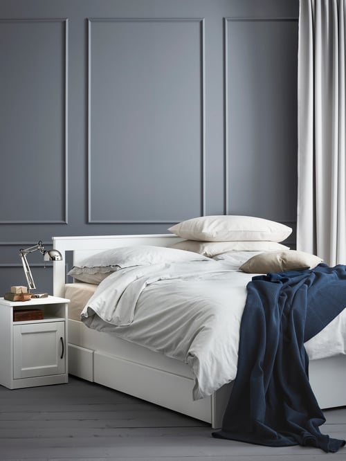 Bedroom with bed frame and mattress with multiple cushions, a white duvet and a bedspread, with a bedside table on the left.
