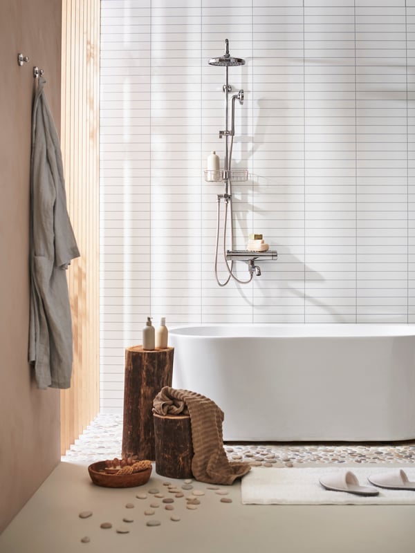 Eine VOXNAN Kombidusche mit Mischbatterie und Duschablage hängt über einer modernen Badewanne. Im Vordergrund hängt ein Bademantel an der Wand und auf dem Kieselsteinboden stehen 2 unterschiedlich hohe Baumstämme auf denen Handtücher und Duschgelflaschen. 