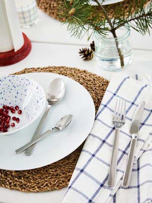 An IKEA 365+ plate on a natural fiber placemat. A white bowl with pomegranate seeds on the plate together with two spoons.