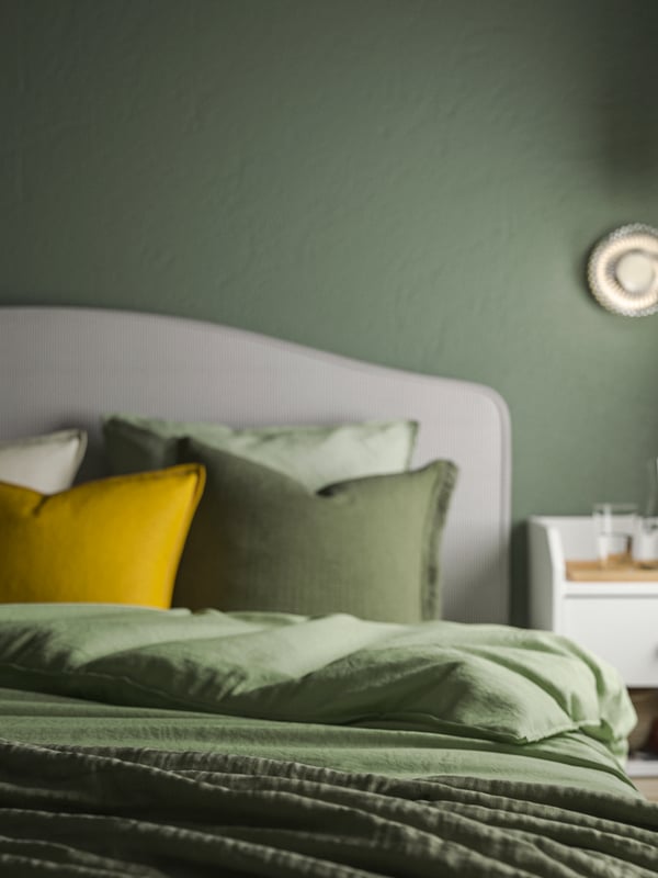 Close-up of upholstered bed with green ÄNGSLILJA duvet cover and pillowcases, with one bright yellow pillow mixed in.