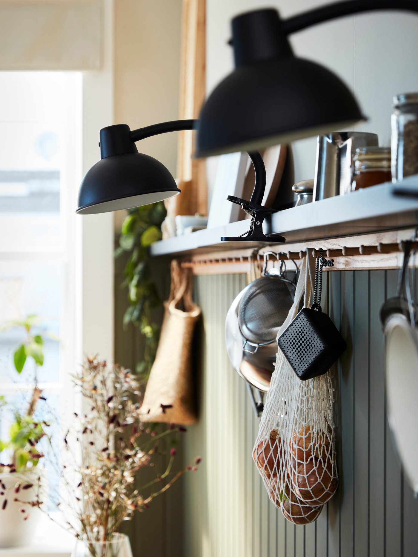 Två SKURUP klämspotar är fastsatta på en bildlist ovanför en VAPPEBY portabel Bluetooth-högtalare som hänger från en knopp.