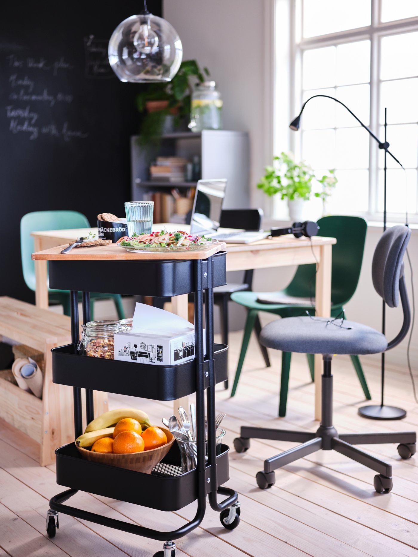 A black RÅSKOG trolley with food and dining accessories stands by an INGO pine table. Above the table hangs a JAKOBSBYN lamp.