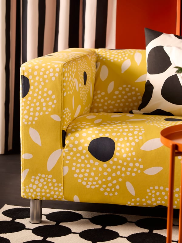 A white and yellow floral-patterned KLIPPAN cover for 2-seat sofa on a sofa sitting on a dotted black and white rug.