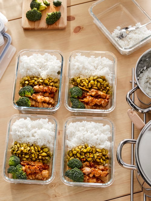 Close up view of kitchen with dining table, food containers with home-cooked meals, pots with lids, spoon, chopping board. 