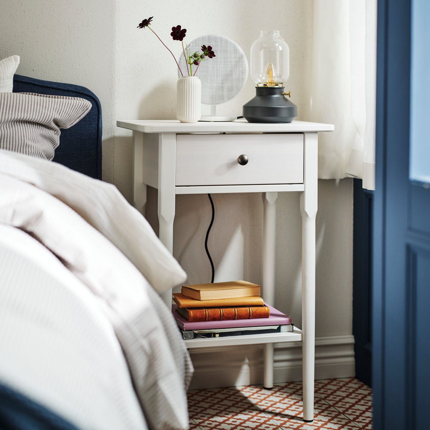 A BLÄSTÅSEN bedside table by a RAMNEFJÄLL bed with a SOLSKYDD Bluetooth speaker on top and some books on the lower shelf.
