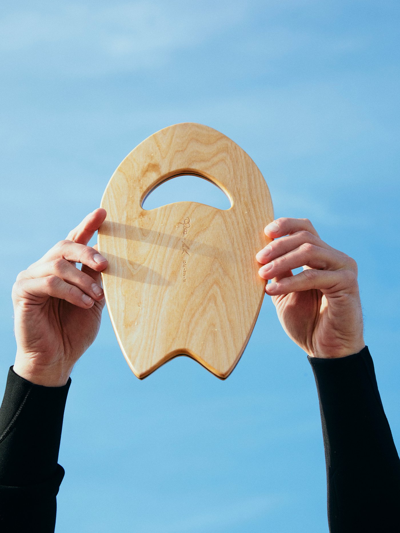 목재 KÅSEBERGA 코세베리아 핸드플레인을 들고 푸른 하늘을 향해 서핑을 하고 있는 블랙 서핑슈트를 입은 서퍼의 두 팔.