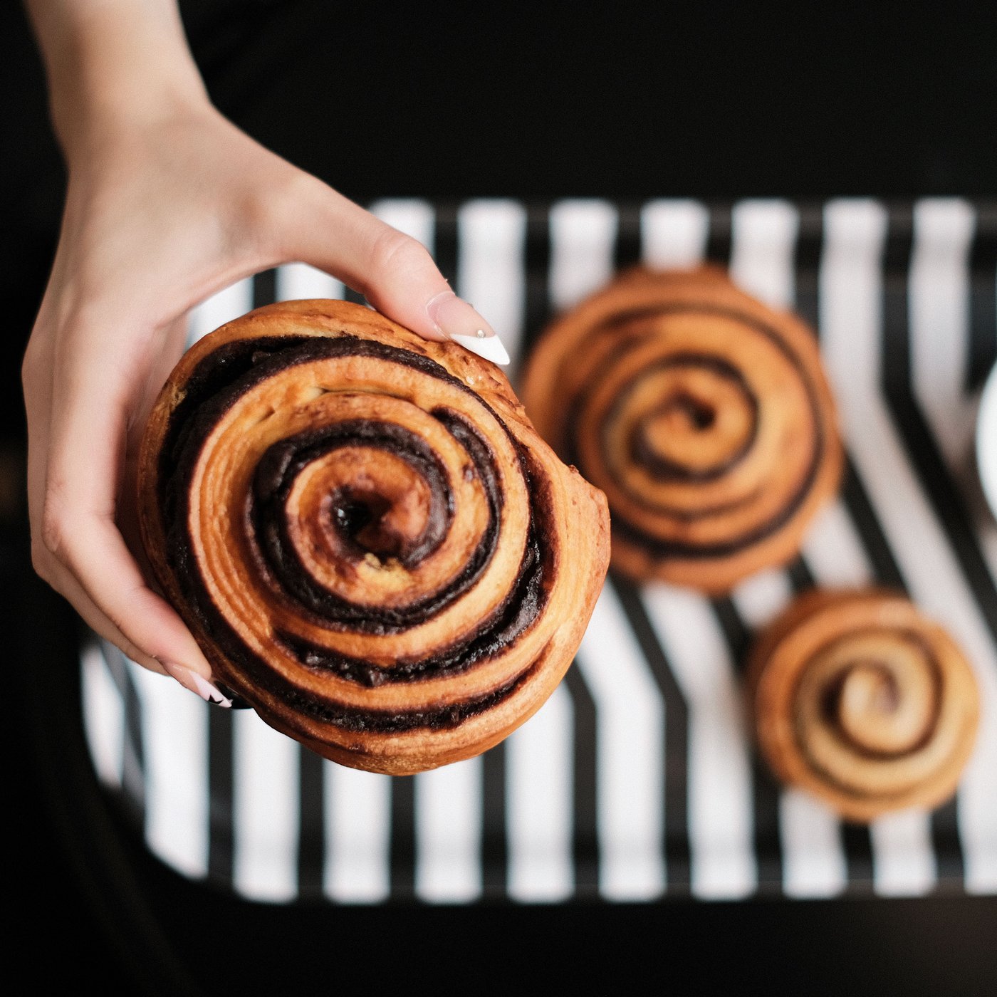 Eine Hand hält eine Schokoladenschnecke. Darunter sind zwei weitere Schnecken auf einem schwarz-weiß gestreiften Tablett zu sehen,