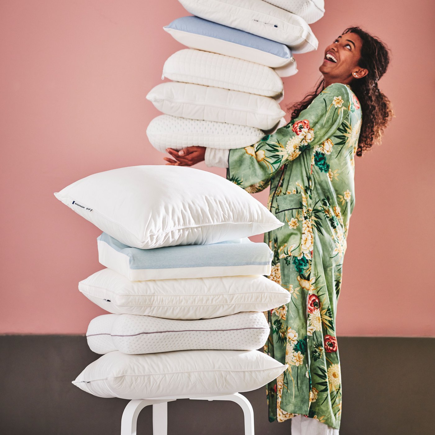 A person holds a high pile of various ergonomic pillows. A smaller pile is in front with a SKOGSFRÄKEN pillow on top.