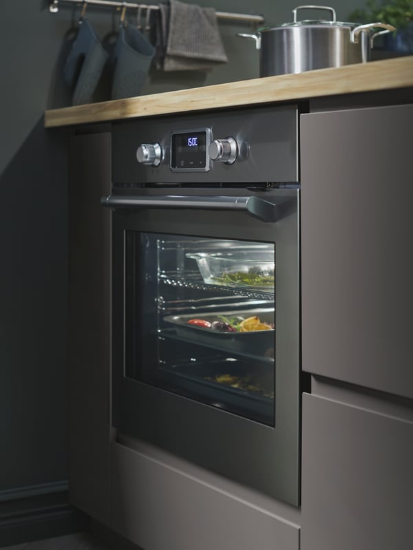 Stainless steel SMAKSAK oven in a kitchen with dark grey VOXTORP fronts, ash/veneer worktop and a rail with hooks.