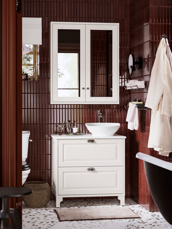 A bathroom with dark tiled walls featuring a TÄNNFORSEN wash-stand with drawers, white basin, and hanging towels.