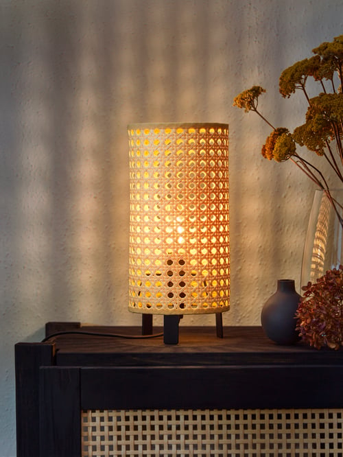 Dimly lit living room area with a focused shot of a lit table lamp placed on a wooden surface, and a vase to the side.