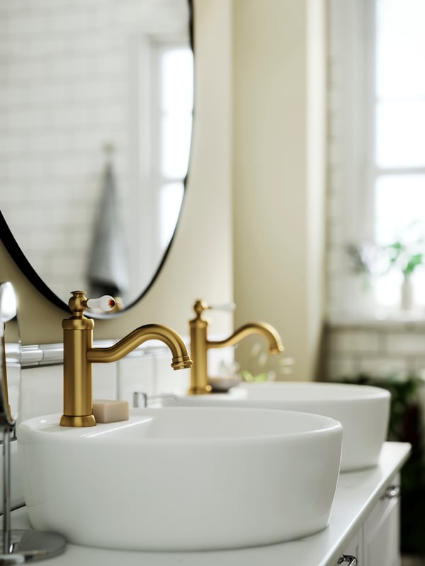 Two HAMNSKÄR wash-basin mixer taps in brass colour on white basins with a round mirror and light decor.
