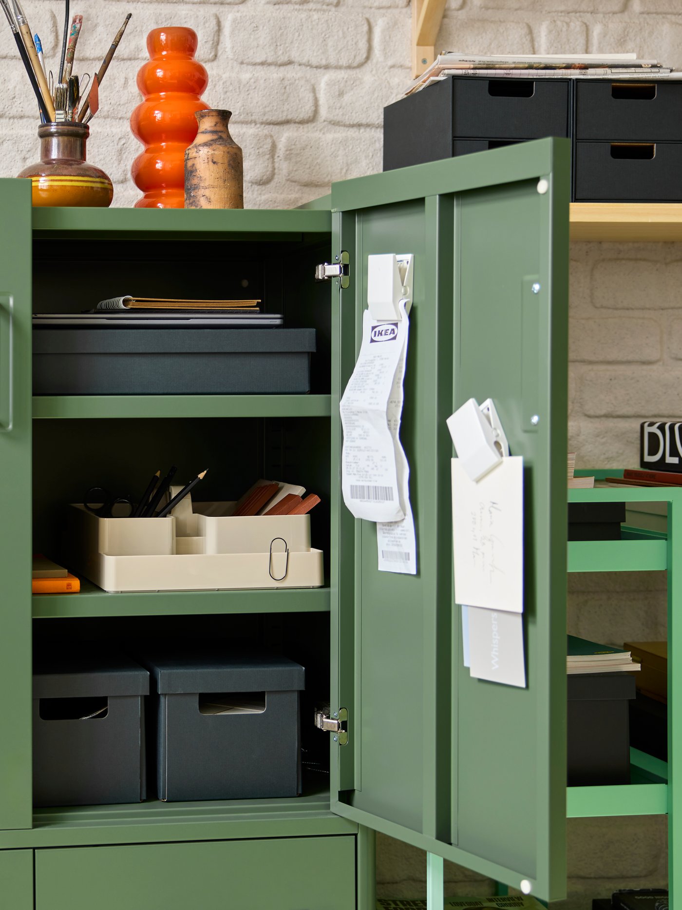 An IDÅSEN cabinet with one door open, showing anthracite paper boxes and a grey-beige KUGGIS desk organiser set.