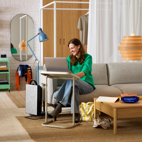 A person sitting on a light grey sofa, working on a beige BJÖRKÅSEN laptop stand with an air purifier next to it.