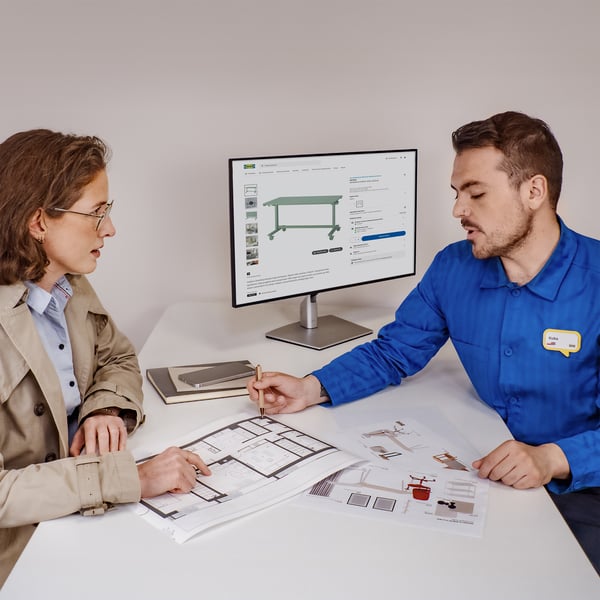 Projektant IKEA siedzący przy biurku w niebieskiej koszulce i omawiający z klientką biznesową rozwiązania do biura. Przy biurku siedzi również klientka słuchający o rozwiązaniu i wskazująca na plan biura.