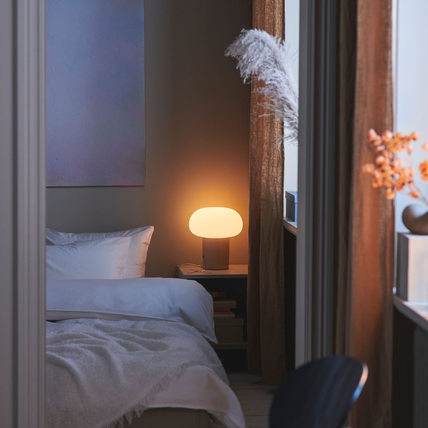 Ett sovrum med en bäddad säng och en varmglödande bordslampa på sängbordet bredvid, med långa gardiner vid fönstret.