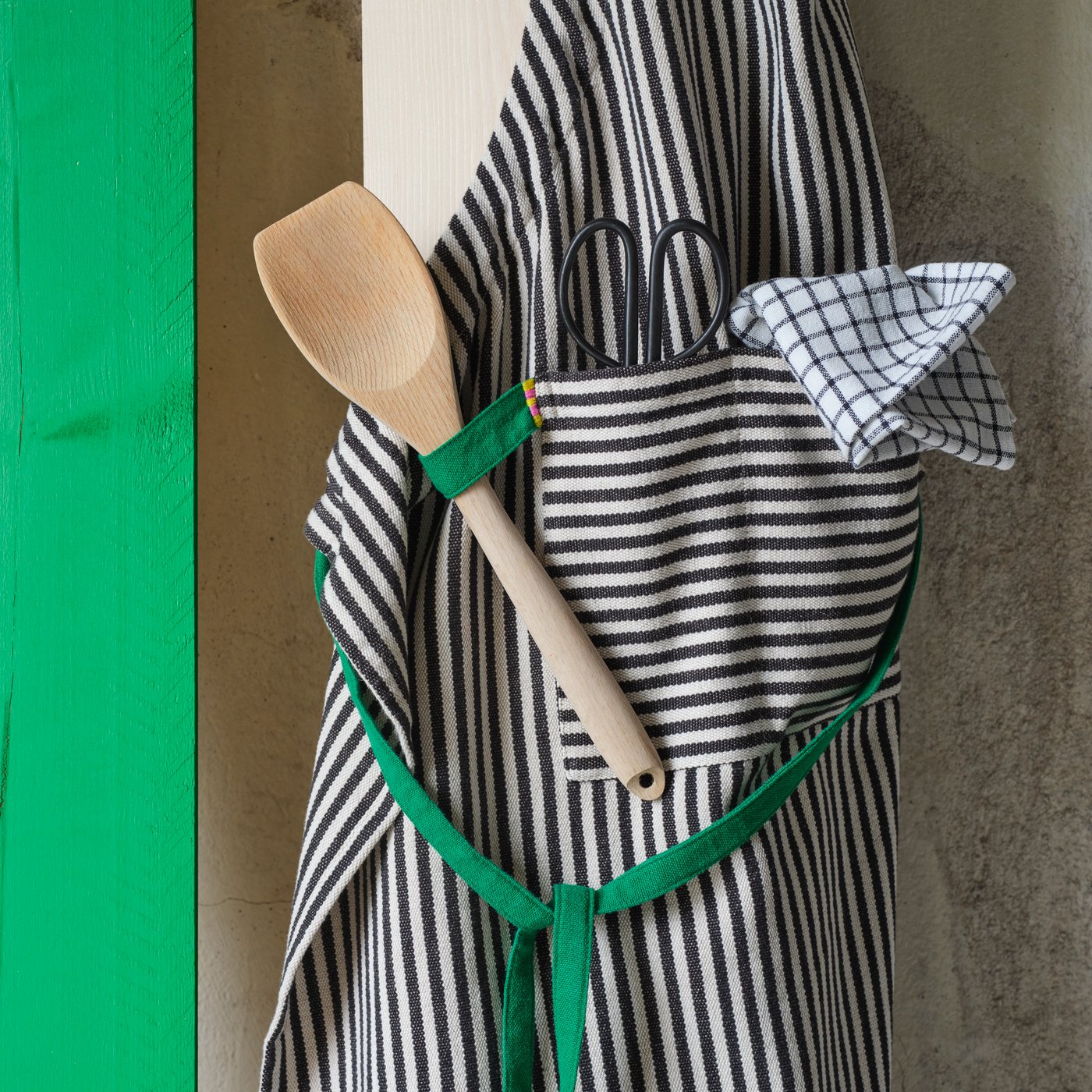 A striped MÄVINN apron is hanging on a wooden plank. The green loop holds a spatula and the front pocket a pair of scissors.