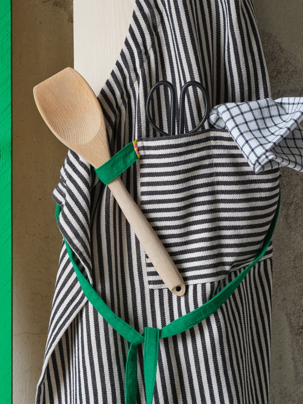 A striped MÄVINN apron is hanging on a wooden plank. The green loop holds a spatula and the front pocket a pair of scissors.