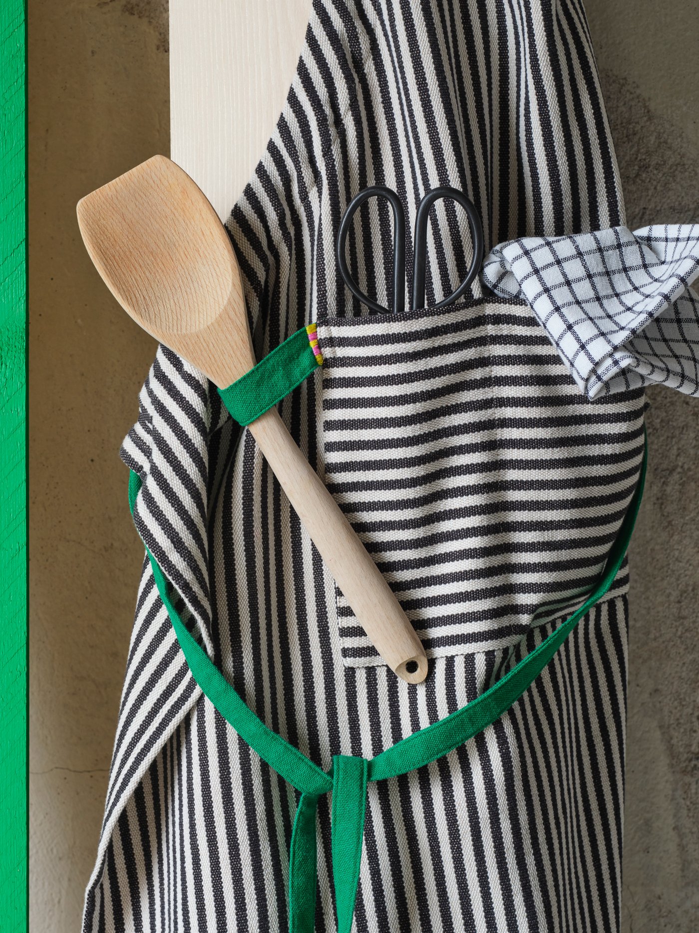 A striped MÄVINN apron is hanging on a wooden plank. The green loop holds a spatula and the front pocket a pair of scissors.