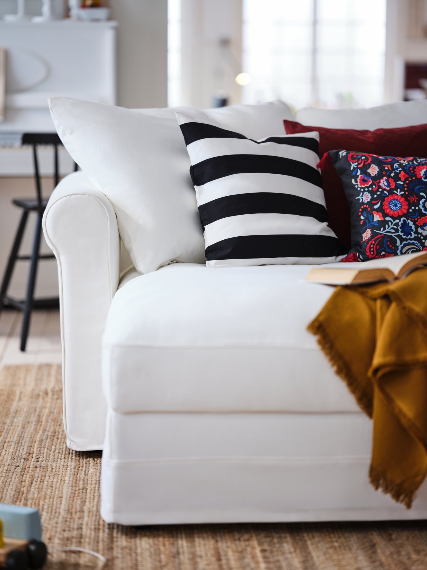 A living room where various colourful cushions and a throw occupy the chaise longue part of a white GRÖNLID four-seat sofa.