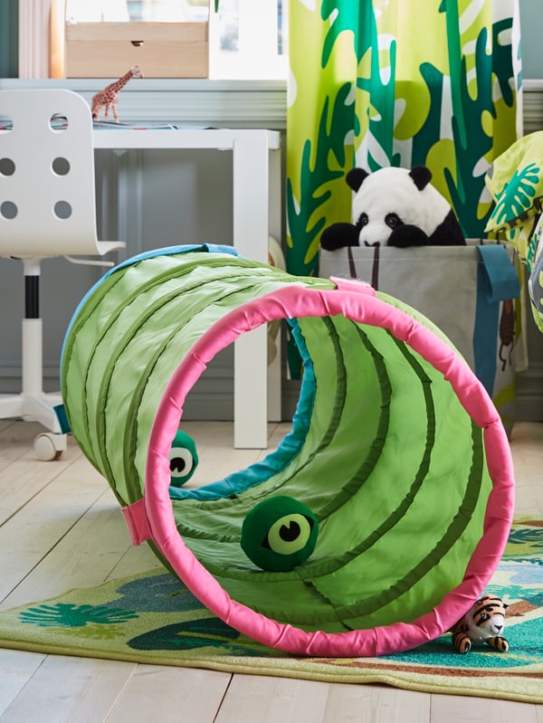 A children’s room where a green BUSA play tunnel lies on a DJUNGELSKOG low pile rug near a desk under a window.