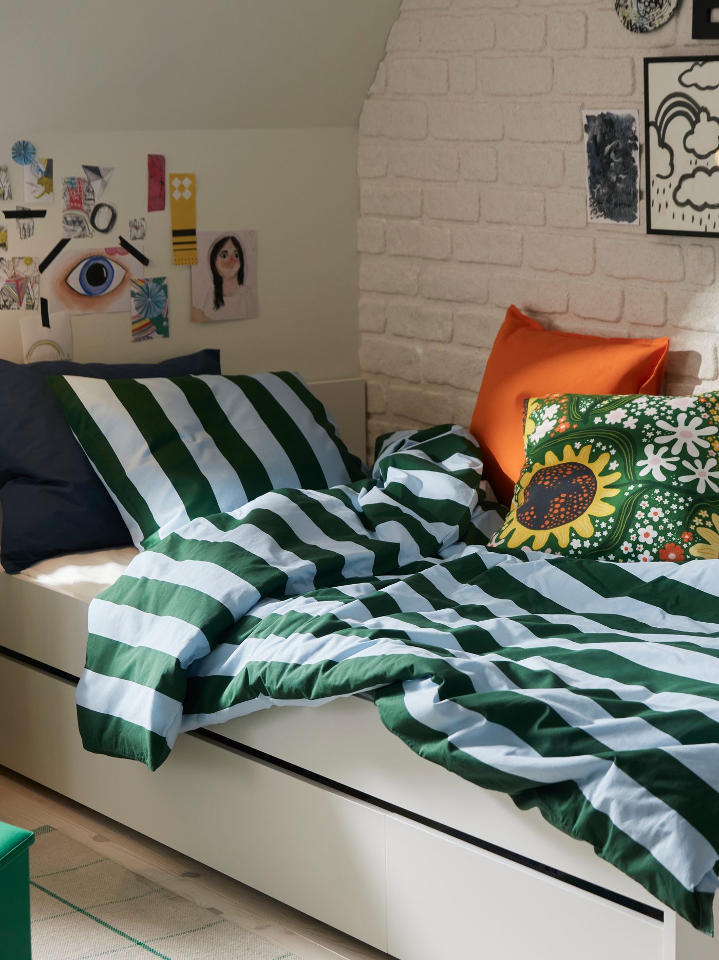 A bedroom with dark green and light blue striped SLÅNHÖSTMAL bedlinen on a white bed by a white brick wall.