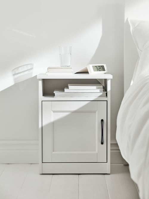 A SONGESAND bedside table in white with books, a glass of water and an alarm clock is by a bed with white bed linen.