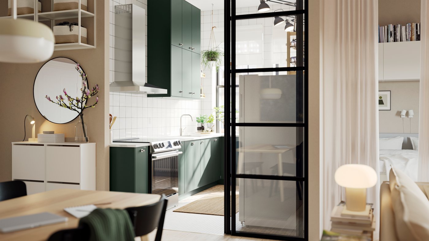 An open kitchen with deep green HAVSTORP kitchen fronts and wall cabinets up to the ceiling, next to a bright living room.