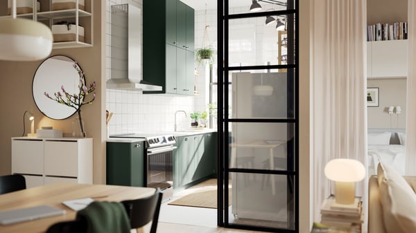 An open kitchen with deep green HAVSTORP kitchen fronts and wall cabinets up to the ceiling, next to a bright living room.