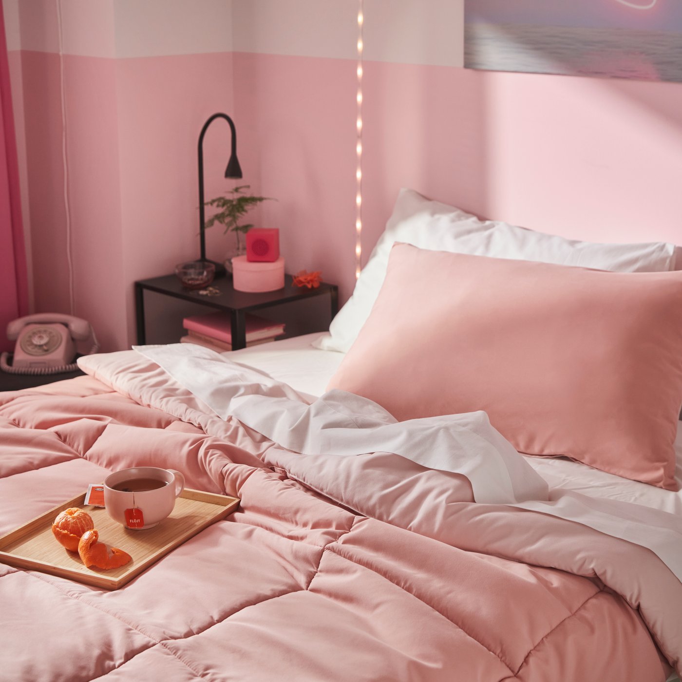 A bedroom styled with a pink HEDFRYLE comforter set draped over a bed, accompanied by a tray holding tea and fruit.