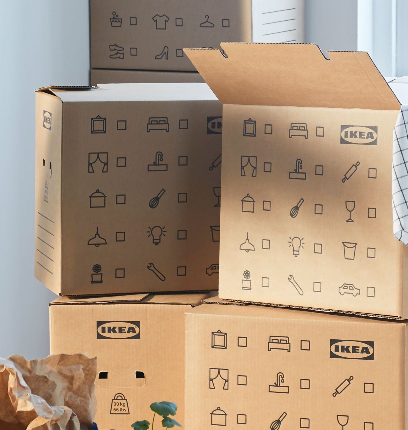 Stack of DUNDERGUBBE moving boxes in corner of a room with window plus SOCKERBIT storage boxes, FRAKTA bag and plants.