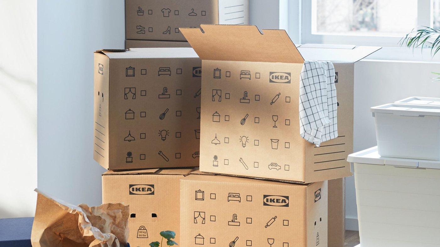 Stack of DUNDERGUBBE moving boxes in corner of a room with window plus SOCKERBIT storage boxes, FRAKTA bag and plants.