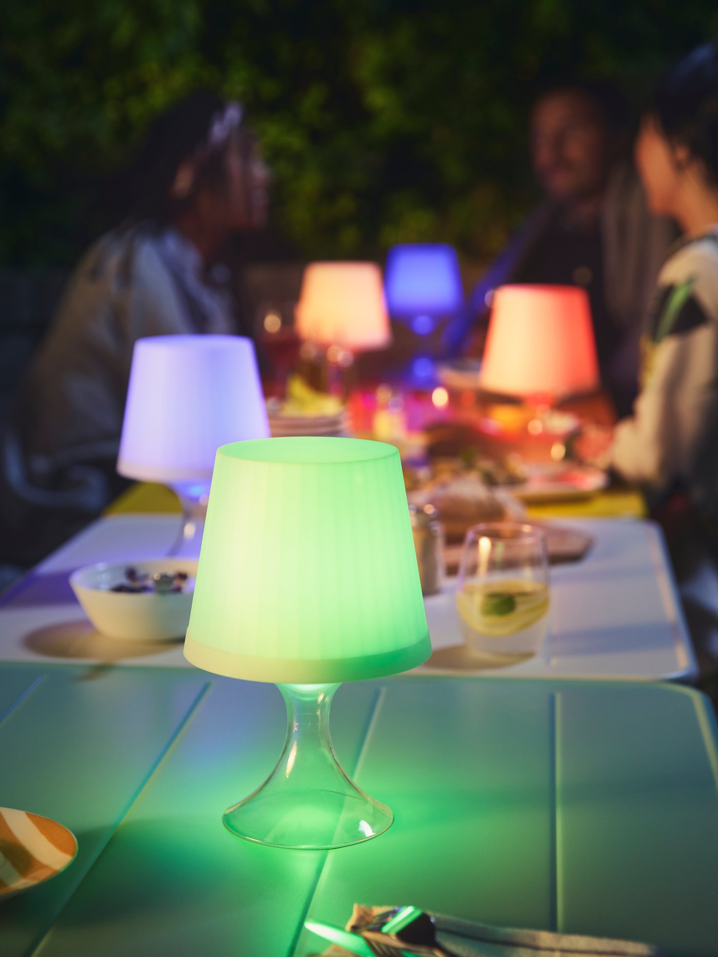 An outdoor seating area is set with LAMPAN lamps in blue, red, and green, lighting up an evening chat.
