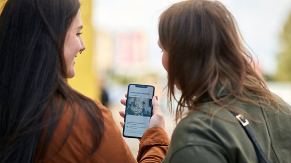 屋外で2人がスマートフォンを見ている様子。スマートフォンの画面にはテキストと画像が表示されており、背景にはぼんやりとした建物や木々が見える。