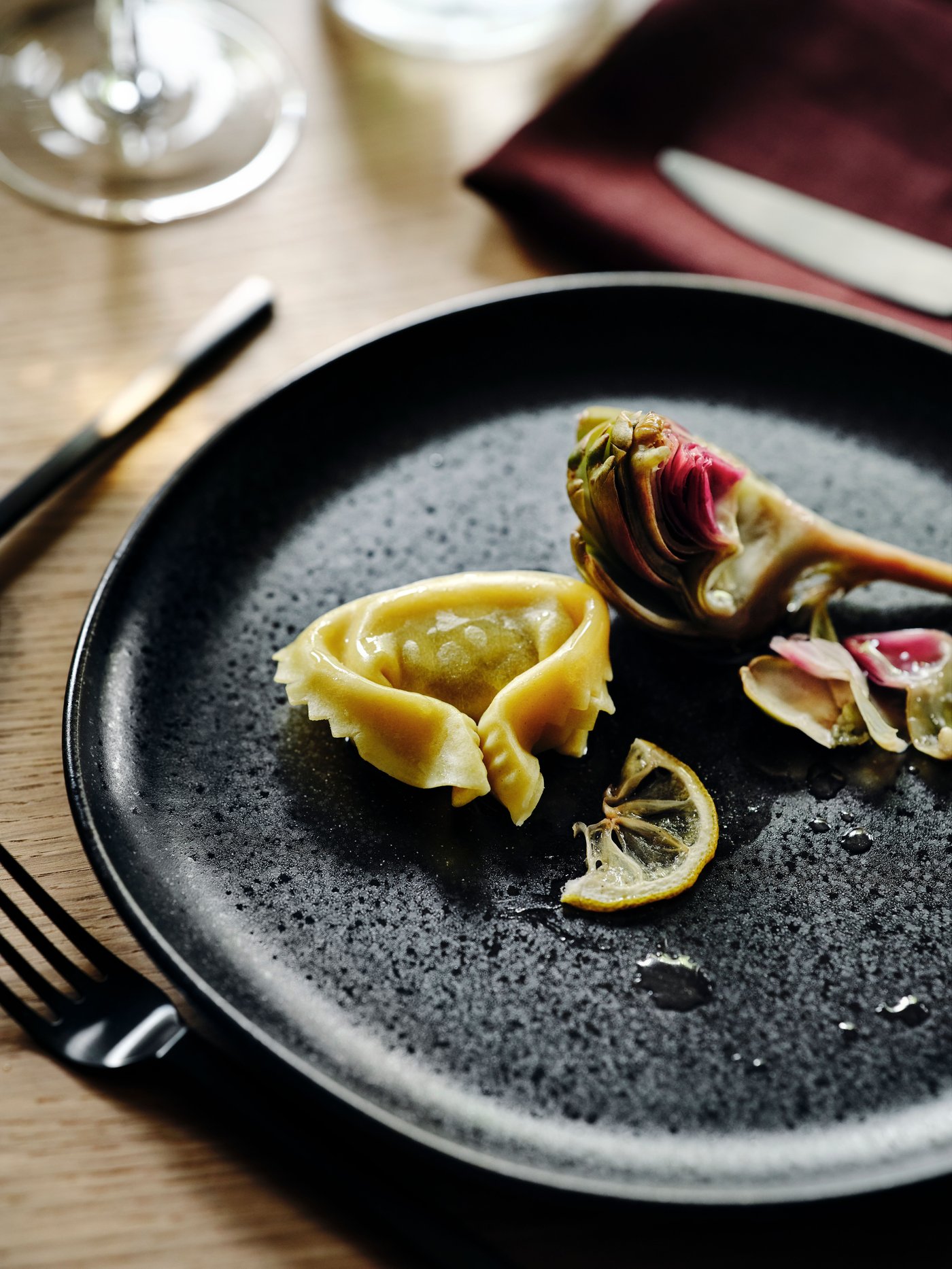 A black STOCKHOLM 2025 plate with food on it sitting on a tabletop, black cutlery and a dark red napkin next to it.