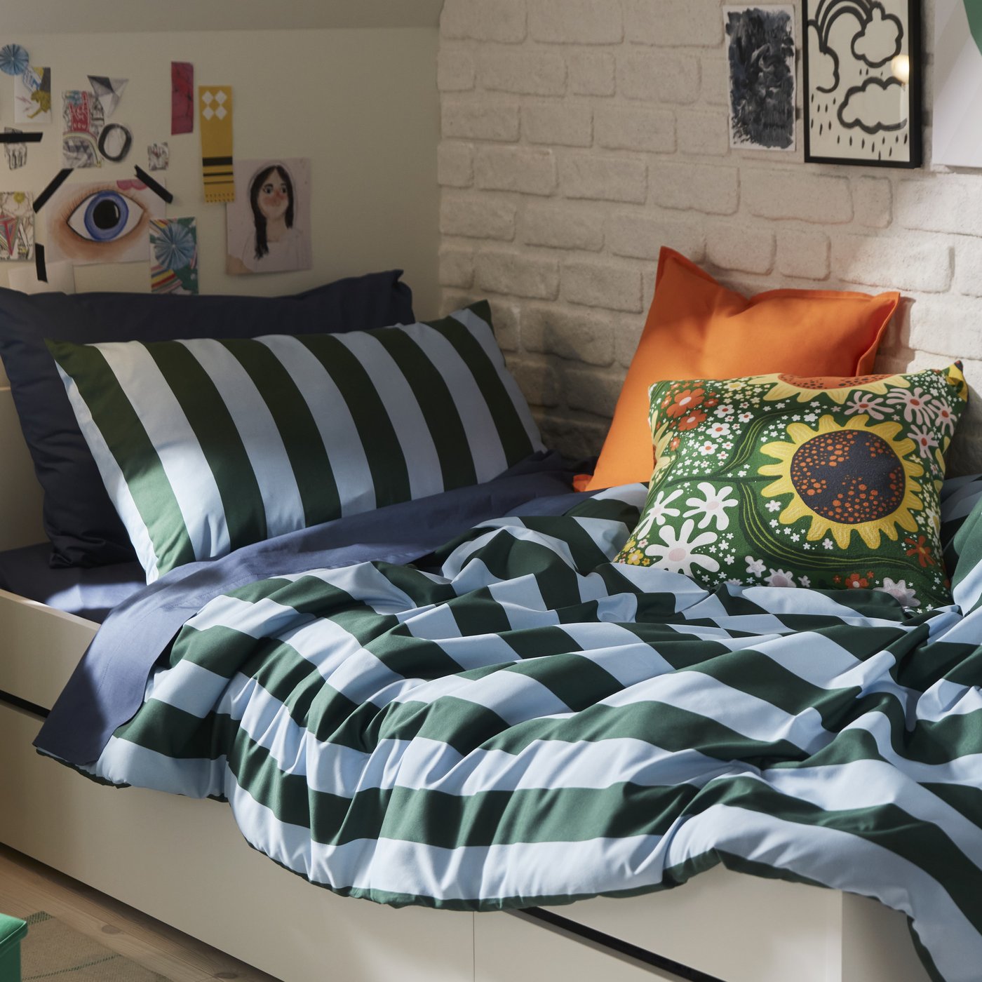 A bedroom with dark green and light blue striped SLÅNHÖSTMAL bedlinen on a white bed by a white brick wall.