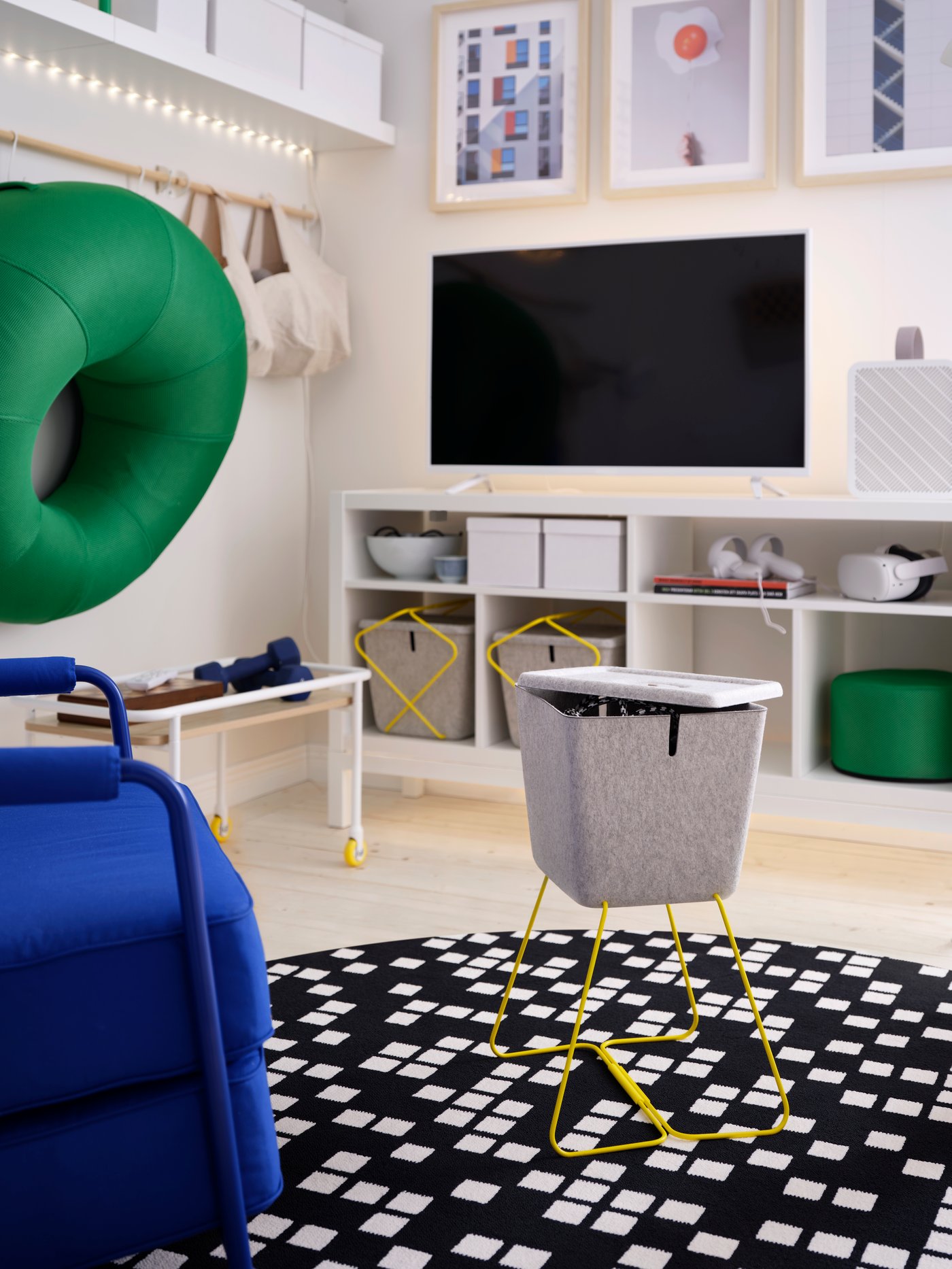 A grey, felt BRÄNNBOLL basket with yellow legs stands as a side table on a checkered rug opposite a console gaming TV setup.