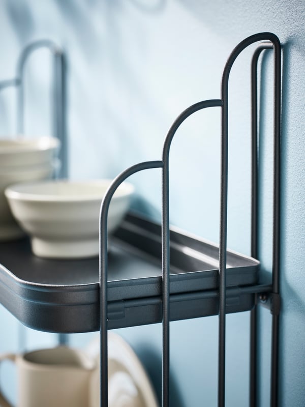 A black APOTEKARE wall shelf with two levels, mounted on a light blue wall. It’s holding white plates and a cup.