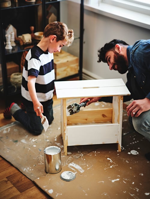 A child is kneeling next to an adult who is painting a PERJOHAN stool with storage. Next to the stool is a can of paint.