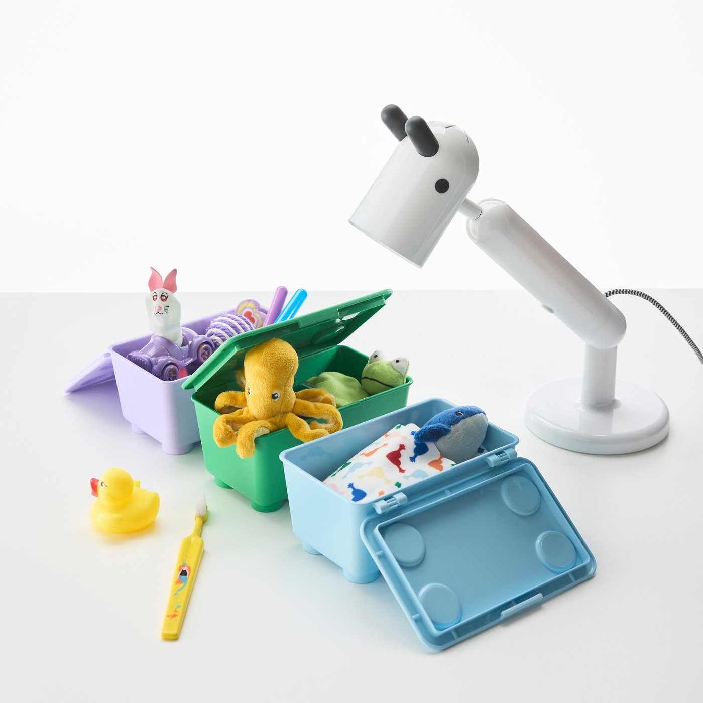 Three GLIS storage boxes with lid, in lilac, green, and pink, filled up with toys tucked in for bed.