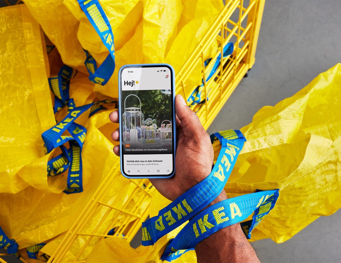 Eine Person hält Handy mit einer geöffneten IKEA App im IKEA Einrichtungshaus vor gelben FRAKTA Taschen