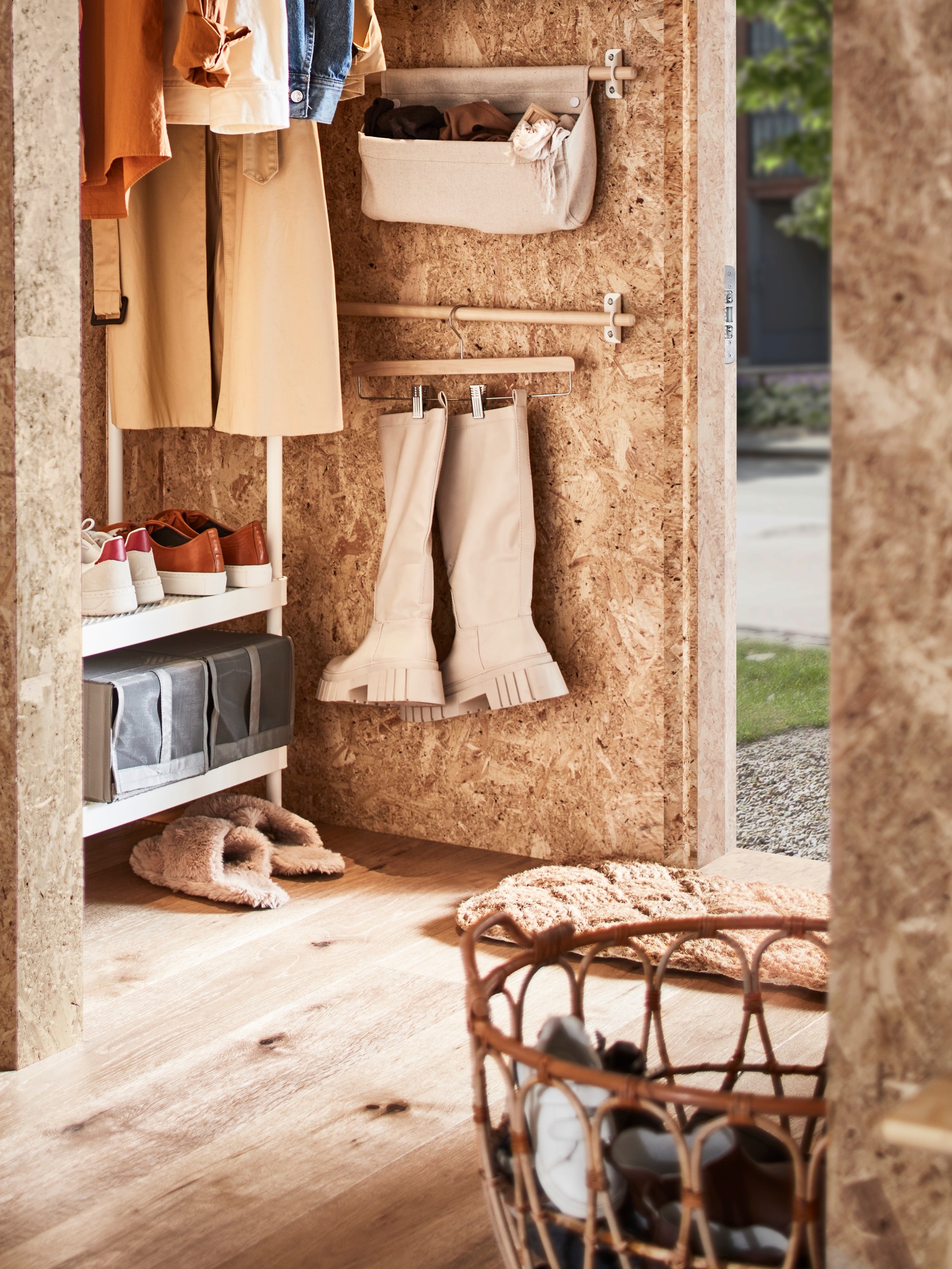 Un hol cu un suport de haine alb cu spațiu de depozitare pentru pantofi, un coș din ratan cu pantofi, plus o șină cu cizme pe un umeraș pentru fuste.