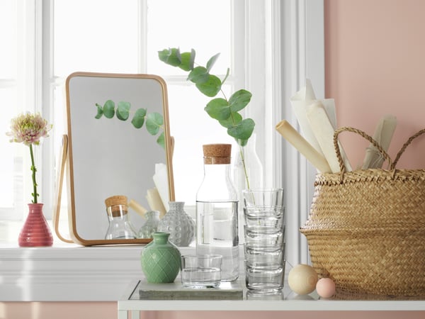 A collection of home decor items including a mirror, small vases, a glass carafe with glasses, and a basket all sitting by a window on a pink wall.