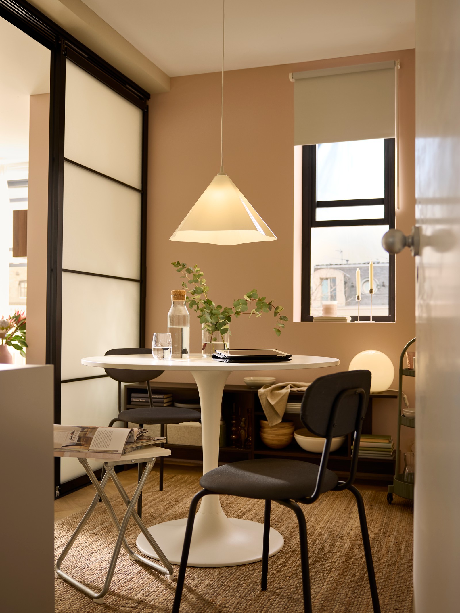 Black ÖSTANÖ chairs and a white GUNDE folding stool surrounding a white DOCKSTA table in a dining area with sliding doors.