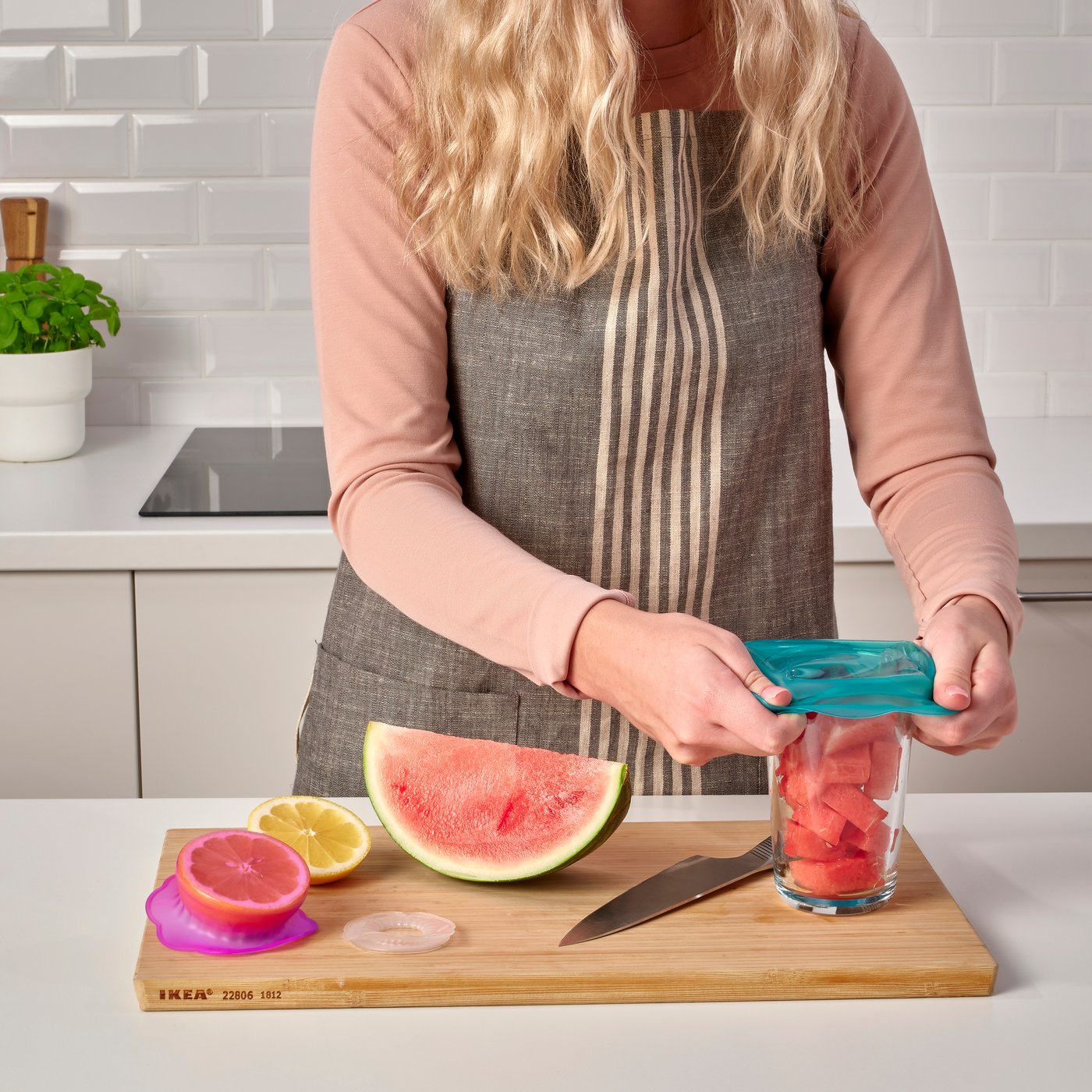 Una persona col·loca una campana d'aliments de silicona ÖVERMÄTT de color turquesa sobre un got amb síndria trossejada damunt d'una fusta de tallar.